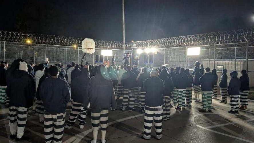 Group of people in striped clothing gathered in a dimly lit fenced area at night.