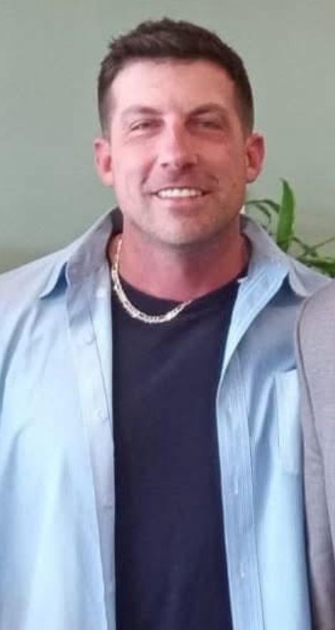 Man wearing a light blue button-up shirt over a black t-shirt with a silver chain necklace.