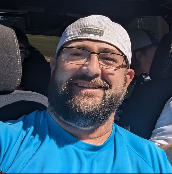 Smiling man wearing a white cap and glasses in a car, with a bright blue shirt.