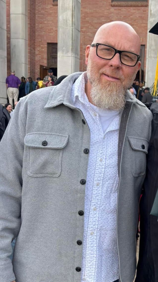 Man with glasses and beard wearing a gray jacket at an outdoor event.